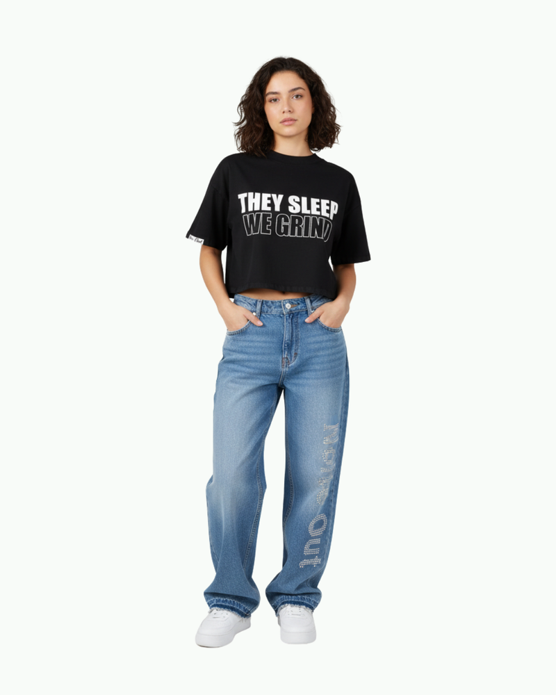 Female model wearing a black cropped ‘They Sleep We Grind’ graphic tee paired with Nite Owl rhinestone-detail blue denim jeans, showcasing modern women’s streetwear essentials, relaxed-fit jeans, motivational crop tops, and elevated casual outfits for shoppers searching for trendy luxury streetwear looks.